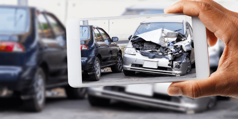 A smartphone captures a photo of a heavily damaged silver car in an urban area, surrounded by other vehicles. The foreground shows a hand holding the phone.