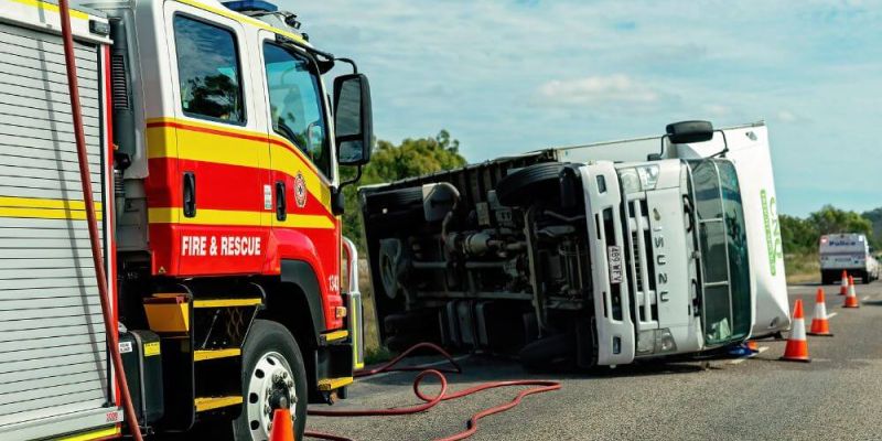 A fire and rescue truck assists near an overturned Isuzu truck on a road. Traffic cones surround the accident scene. Other emergency vehicles are visible in the background.