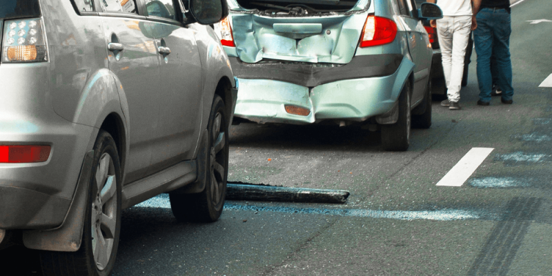 Two damaged cars are stopped on a highway, with their rears visibly dented. Two people, standing nearby, survey the accident scene, one pointing towards the damage.
