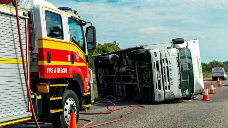 A fire and rescue truck assists near an overturned Isuzu truck on a road. Traffic cones surround the accident scene. Other emergency vehicles are visible in the background.
