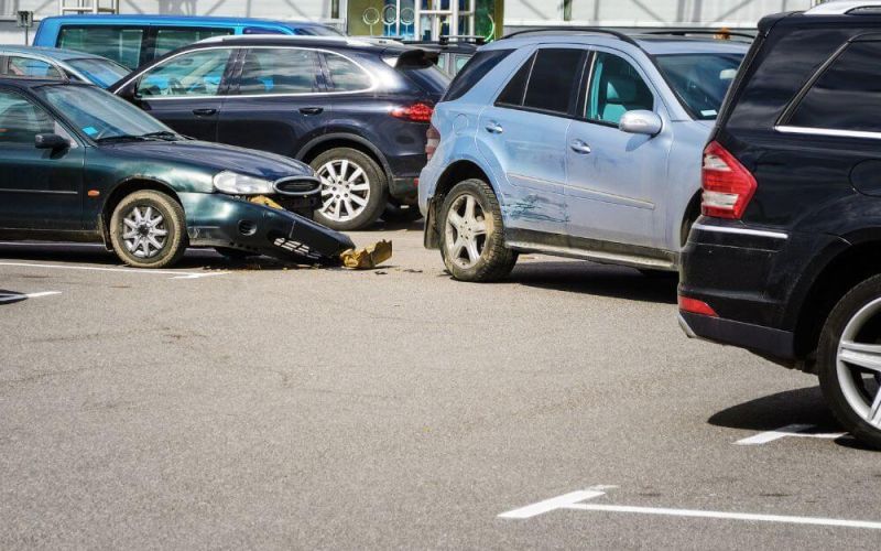 A green car collides with a blue SUV in a parking lot. The SUV's rear is dented, and the green car's front bumper is damaged. Surrounding parked cars create a busy environment.