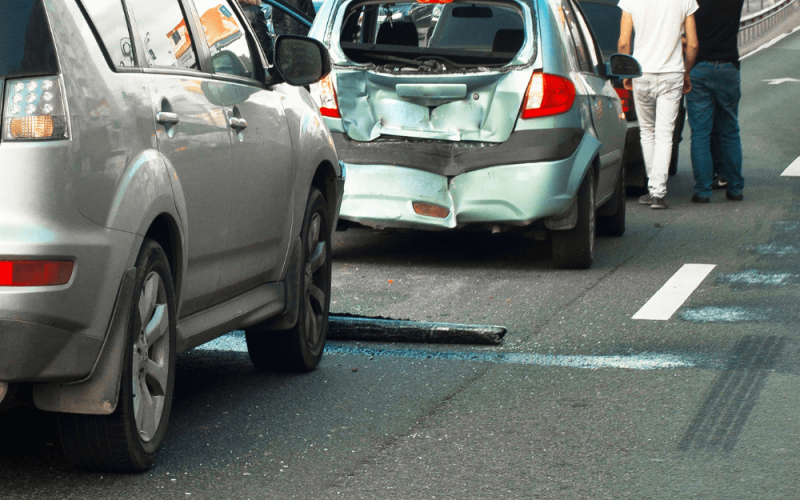 Two damaged cars are stopped on a highway, with their rears visibly dented. Two people, standing nearby, survey the accident scene, one pointing towards the damage.
