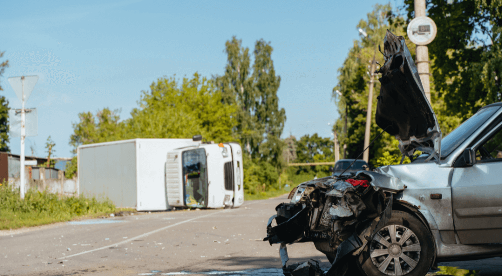 A damaged silver car sits on a road, its front crumpled and hood raised. An overturned white truck lies nearby, surrounded by debris, under a clear blue sky with greenery in the background.