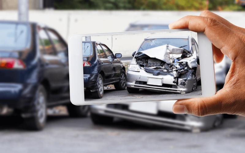 A smartphone captures a photo of a heavily damaged silver car in an urban area, surrounded by other vehicles. The foreground shows a hand holding the phone.