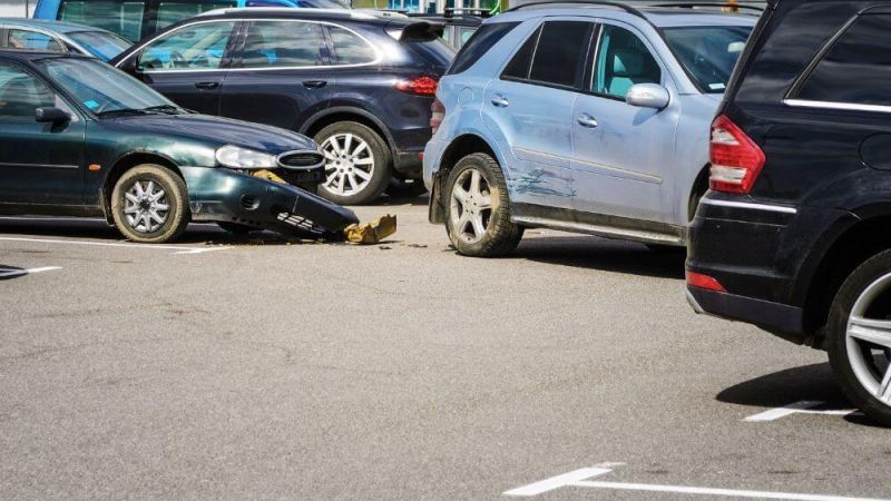 A green car collides with a blue SUV in a parking lot. The SUV's rear is dented, and the green car's front bumper is damaged. Surrounding parked cars create a busy environment.