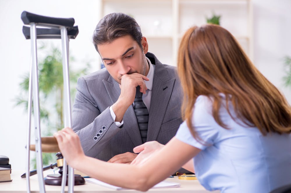 Why Miami Residents Choose Klotzman Law Firm as Their Personal Injury Lawyer – Klotzman Law Firm A man in a suit, contemplating with hand on chin, listens to a woman across a desk. Nearby are crutches and a gavel, suggesting a legal or medical consultation.