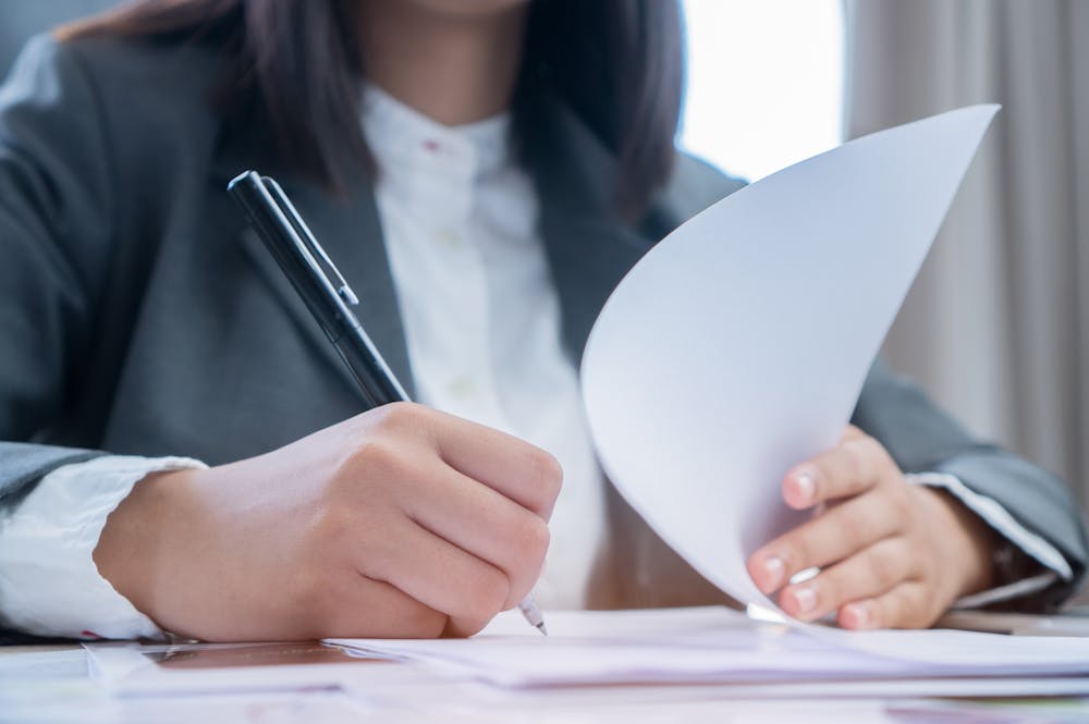 How Can I File a Claim for Hurricane Damage in Orlando – Klotzman Law Firm A person in a suit is writing on a piece of paper with a pen while holding another sheet of paper, sitting at a desk with natural light coming from a nearby window.