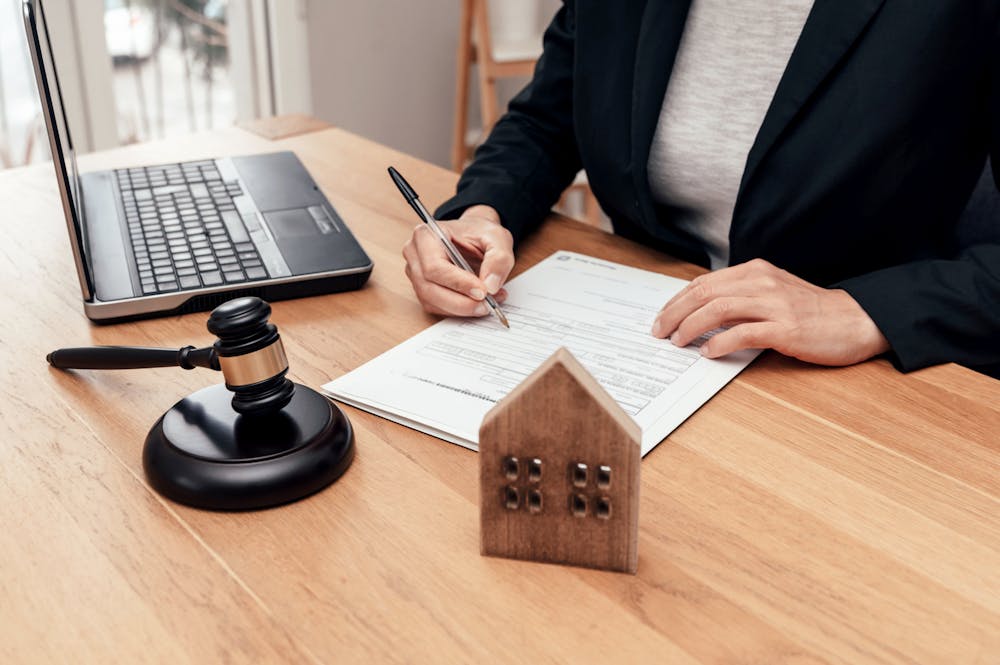 How Our Sarasota Hurricane Damage Lawyer Can Help – Klotzman Law Firm A person in a suit writes on a document at a wooden desk, beside a laptop and a gavel, with a small wooden house model in the foreground, suggesting a legal or real estate context.