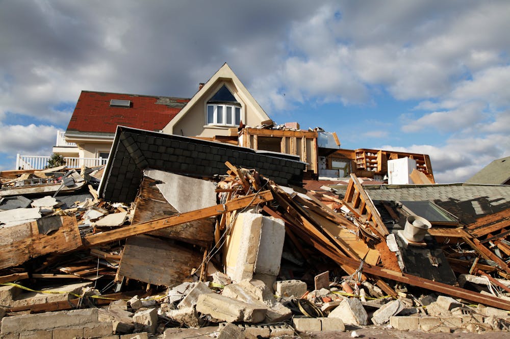 Types of Hurricane and Tropical Storm Damage Claims We Handle in Orlando – Klotzman Law Firm A collapsed structure, comprising debris and scattered building materials, is foregrounded against a partially intact house. The scene is set under a sky with scattered clouds, indicating recovery challenges.