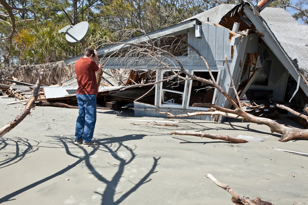 What Should I Do After My Property Sustains Hurricane Damage – Klotzman Law Firm A man stands on a sandy surface, photographing a damaged house with debris and uprooted branches scattered around, indicating a recent disaster in a wooded area.
