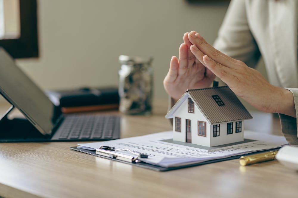 What Types of Property Damage Are Covered Under Homeowners Insurance – Klotzman Law Firm A person is placing their hands above a small model house on a desk, surrounded by paperwork and a tablet, suggesting a focus on real estate or insurance in an office setting.
